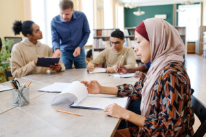 Muslim female immigrant sitting at table looking through sheets of paper with grammar tasks during English lesson